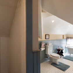 Cozy bathroom with a sloped ceiling, featuring a freestanding bathtub, a toilet, and a vintage-style sink. The space has hexagonal floor tiles, blue wall paneling, and two windows with sheer curtains.