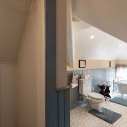 Cozy bathroom with a sloped ceiling, featuring a freestanding bathtub, a toilet, and a vintage-style sink. The space has hexagonal floor tiles, blue wall paneling, and two windows with sheer curtains.