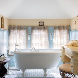 A bathroom with a freestanding clawfoot bathtub, blue and beige walls, hexagonal floor tiles, and a wooden vanity set with a mirror and chair. Various toiletries are placed on a side table beside the tub.